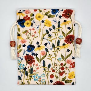 May include: A white drawstring bag with a floral print featuring red, yellow, blue, and green flowers. The bag has white drawstrings with brown wooden beads.
