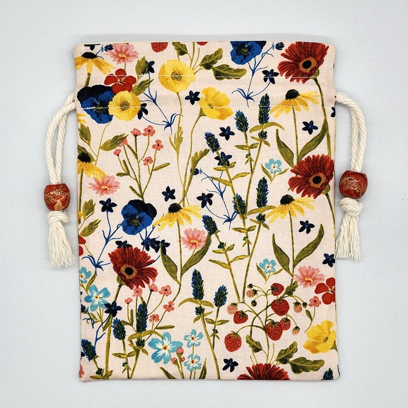 May include: A white drawstring bag with a floral print featuring red, yellow, blue, and green flowers. The bag has white drawstrings with brown wooden beads.