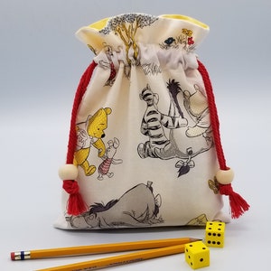 May include: A white drawstring bag with a Winnie the Pooh and friends print. The bag has red drawstrings with wooden beads and is filled with yellow dice.
