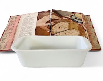 May include: A white rectangular baking dish sits in front of an open cookbook. The cookbook shows a picture of sliced bread and a recipe. The cookbook is bound with metal rings and has red and cream-colored pages.