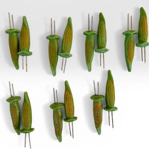 May include: A collection of corn-shaped corn holders. Each holder is green with a yellow-orange corn design and metal prongs. The holders are arranged on a white background.
