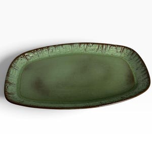 May include: An oval ceramic platter featuring a mottled sage green center and a dark brown rim. The border is decorated with a series of repeating, embossed geometric and abstract symbols that encircle the entire edge of the serving dish.