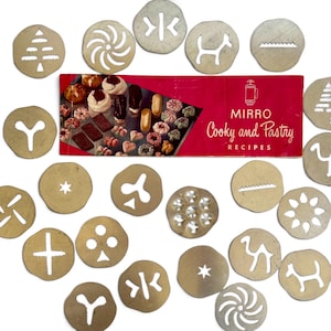 May include: A collection of circular metal cookie press discs featuring various cut-out shapes including stars, animals, and geometric patterns, accompanied by a red Mirro Cooky and Pastry Recipes booklet with images of assorted baked cookies.