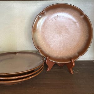 May include: A set of four round ceramic plates with a scalloped edge design. The plates feature a gradient finish that transitions from a deep brown rim to a lighter, speckled tan center. One plate is displayed upright on a brown stand.