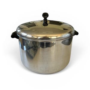 May include: A stainless steel pot with a lid. The pot has a rounded body with two black handles on the sides. The lid has a black knob on top. The pot is sitting on a white surface.