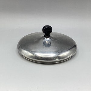 May include: A round, brushed metal lid with a black, rounded handle. The lid has a slightly domed shape and a circular design. The metal surface reflects light, and the handle is centered on top.