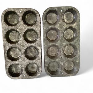 May include: Two vintage, silver-colored muffin tins with a decorative pattern. Each tin has six circular molds with a starburst design in the bottom. The tins are made of metal and are likely used for baking muffins or cupcakes.