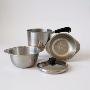 May include: A collection of stainless steel kitchenware, including a small saucepan with a black handle, a strainer, a small bowl, and a lid with a black knob. The saucepan has a copper-colored base.