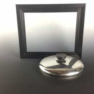 May include: A stainless steel pot lid with a black knob, positioned in front of a black picture frame. The lid reflects the light, and the frame has a simple, rectangular design. The background is a gradient of white and gray.