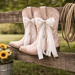 May include: Pale pink cowboy boots with white bows and lace detail, displayed on a wooden fence. A coiled rope hangs beside them. A metal bucket of sunflowers and wildflowers sits in front.