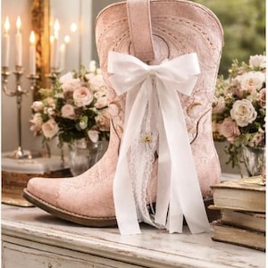 May include: A pale pink cowboy boot with floral embroidery and a white bow. The boot is adorned with white lace and a small gold flower. It's displayed on a white table with a distressed finish, reminiscent of a vintage aesthetic.