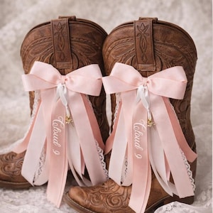 Pode incluir: Um par de botas de cowboy de couro castanho decoradas com la&ccedil;os e fitas cor-de-rosa. As fitas t&ecirc;m o texto "Cloud 9" impresso. As botas est&atilde;o sobre uma superf&iacute;cie de renda branca.