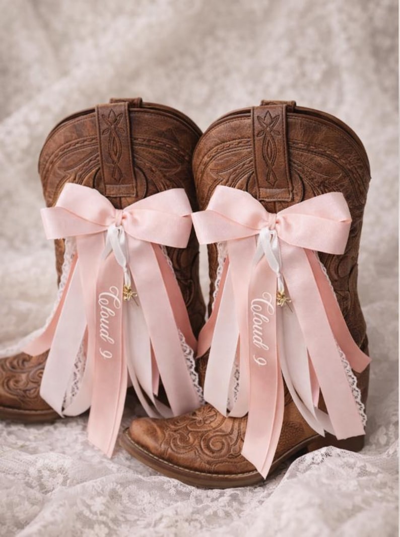 Pode incluir: Um par de botas de cowboy de couro castanho decoradas com la&ccedil;os e fitas cor-de-rosa. As fitas t&ecirc;m o texto "Cloud 9" impresso. As botas est&atilde;o sobre uma superf&iacute;cie de renda branca.