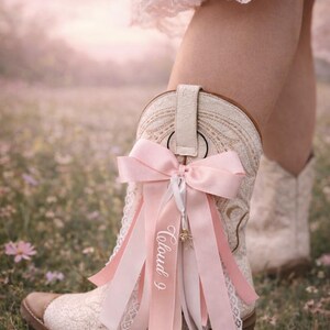 Pode incluir: Bota de cowboy creme com um design floral rendado, adornada com um la&ccedil;o e fitas rosa. As fitas t&ecirc;m o texto "Cloud 9" impresso. A bota tem uma biqueira e um calcanhar de couro castanho.
