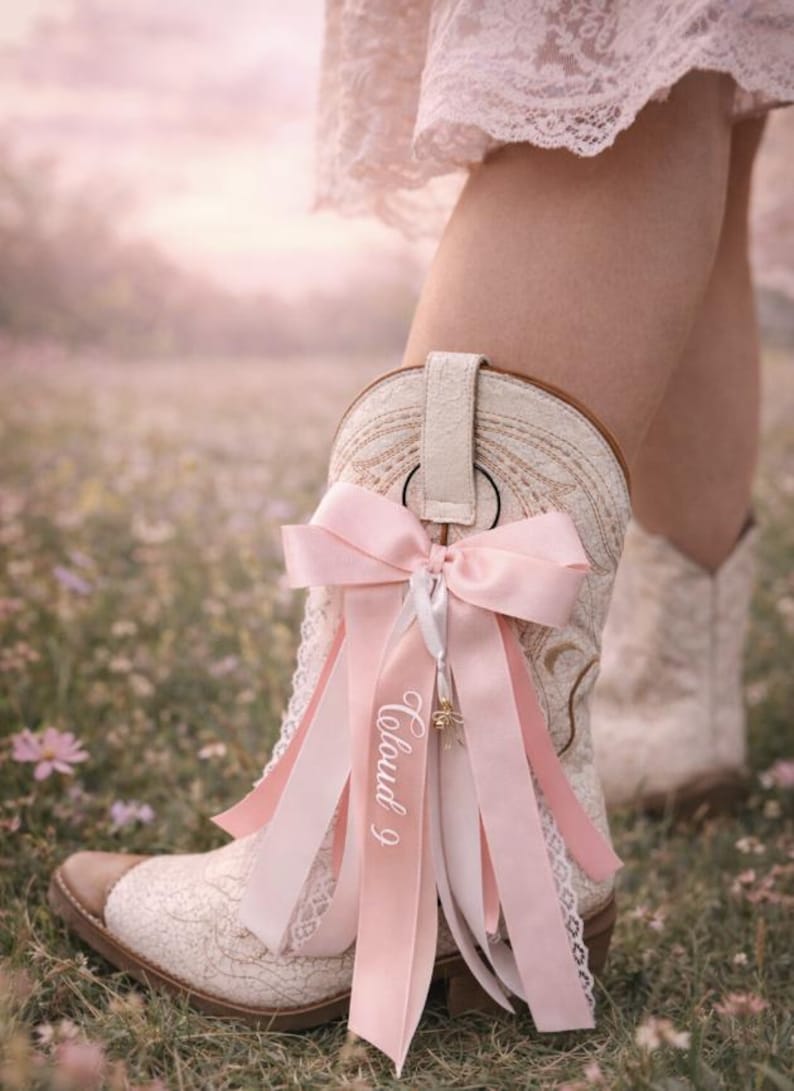 Pode incluir: Bota de cowboy creme com um design floral rendado, adornada com um la&ccedil;o e fitas rosa. As fitas t&ecirc;m o texto "Cloud 9" impresso. A bota tem uma biqueira e um calcanhar de couro castanho.