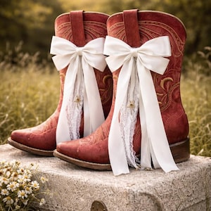 May include: Red cowboy boots with decorative stitching and white bows, set on a vintage suitcase. The boots have white lace detailing. The background is a blurred view of green foliage.