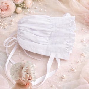 May include: A white baby bonnet with a ruffled edge and tie ribbons. The bonnet is made of a soft fabric and has a gathered design. The background includes pink roses, pearls, and a baby wearing a similar bonnet.