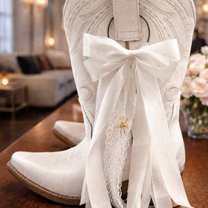 May include: White cowboy boots with detailed stitching and a pointed toe. A large white bow and lace ribbon are tied to the back of one boot, with a small gold charm. The boots are on a wooden surface.
