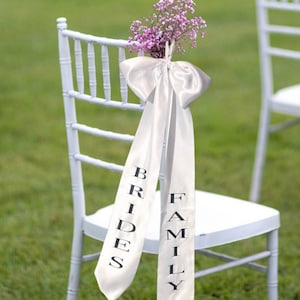 May include: White satin chair sashes with the words "Bride's" and "Family" printed on them. The sashes are tied with a bow and decorated with a bouquet of small pink flowers.