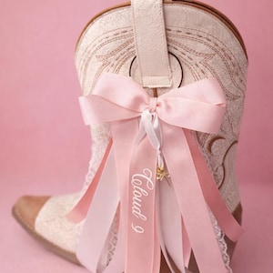 Pode incluir: Uma bota de cowboy creme com costuras intrincadas e biqueira de couro castanho. Um grande la&ccedil;o rosa com fitas a condizer e o texto "Cloud 9" adornam a bota, criando um visual feminino e decorativo.