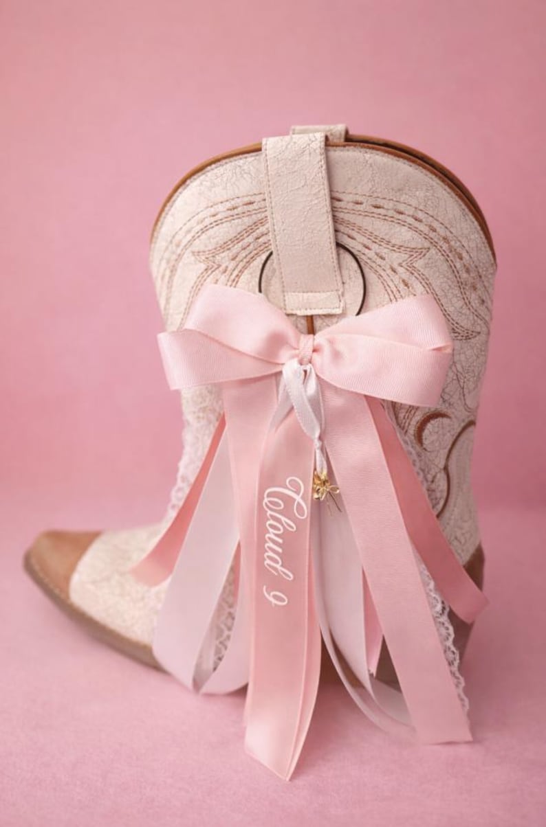 Pode incluir: Uma bota de cowboy creme com costuras intrincadas e biqueira de couro castanho. Um grande la&ccedil;o rosa com fitas a condizer e o texto "Cloud 9" adornam a bota, criando um visual feminino e decorativo.