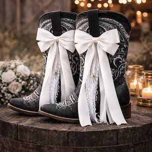 May include: Black cowboy boots with white bows and lace detailing, set on a wooden barrel. The boots have an ornate design. The background includes soft lighting and a bouquet of white flowers, creating a romantic scene.