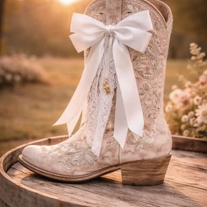 May include: Cream-coloured cowboy boot with detailed embroidery and a white bow. The boot has a pointed toe and a stacked heel, decorated with lace and a small gold charm. It is displayed on a wooden barrel.