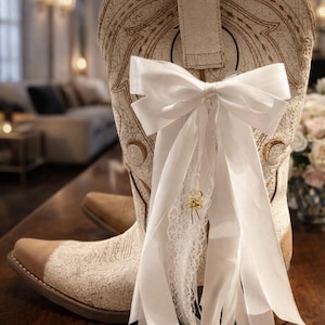 May include: Cream-coloured cowboy boot with a white bow and lace detail. The boot has a textured design and a brown toe. A white bow is tied around the boot's pull strap, with long ribbons and a lace accent.
