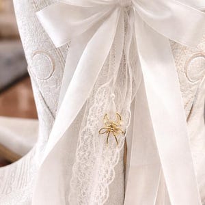 May include: White cowboy boot adorned with a large white satin bow and ribbons. A gold spider charm dangles from a white lace ribbon. The boot has an embossed pattern. A charming detail for a special occasion.