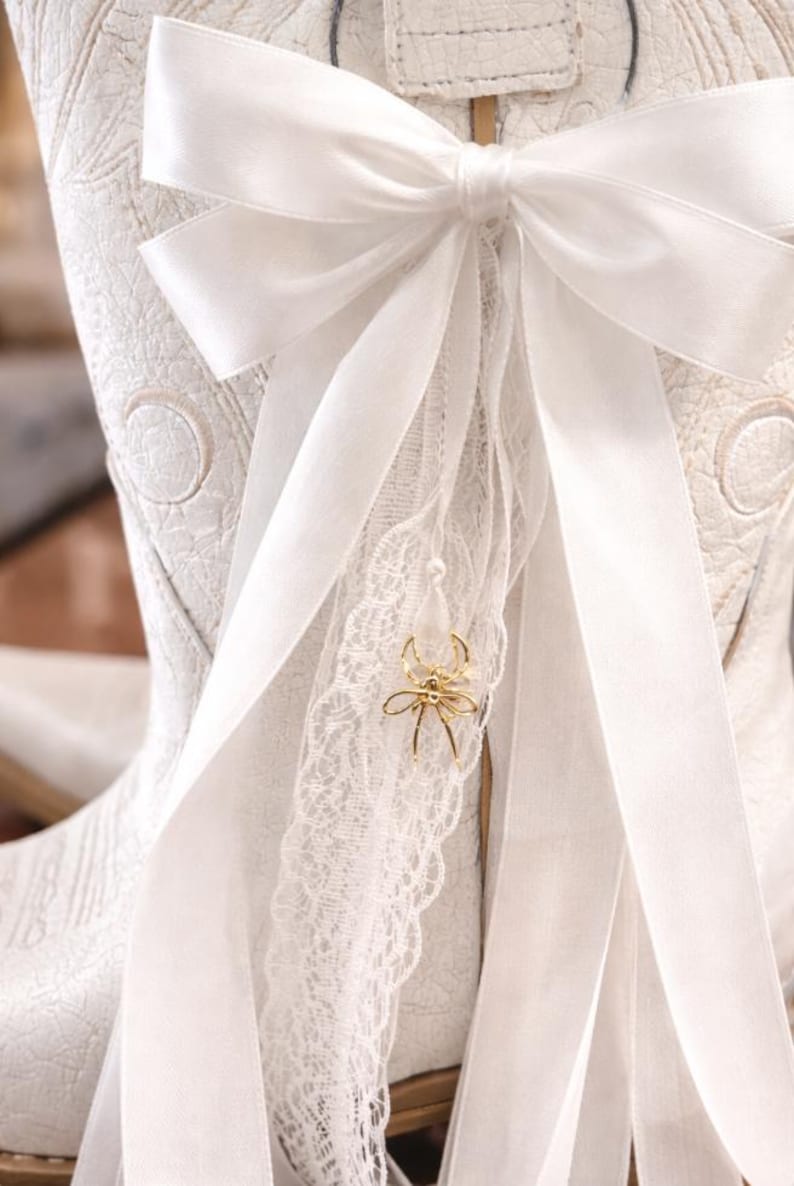 May include: White cowboy boot adorned with a large white satin bow and ribbons. A gold spider charm dangles from a white lace ribbon. The boot has an embossed pattern. A charming detail for a special occasion.
