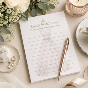Peut inclure: Un bloc-notes blanc avec une illustration légère d'une robe de mariée et le texte Sarah's Bridal Notes Where wedding dreams begin to bloom. Le bloc-notes présente des lignes horizontales et est accompagné d'un stylo or rose, d'une bague et de boucles d'oreilles.
