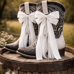 May include: Two black and white cowboy boots with detailed embroidery and white bows. The boots are adorned with white lace and are placed on a wooden barrel, evoking a rustic wedding aesthetic.