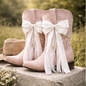 May include: Pale pink cowboy boots with white bows and lace detailing. The boots, with floral embroidery, are displayed on a vintage suitcase. A straw hat is placed nearby. The boots are approximately 30 cm tall.