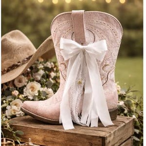 May include: A pale pink cowboy boot with a white bow and lace detail, displayed on a wooden crate. A cowboy hat and flowers are in the background, with soft lighting. The boot is suitable for a wedding or special event.