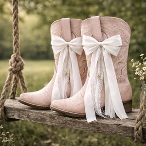 May include: Pale pink cowboy boots with white bows and lace, displayed on a wooden swing. The boots feature floral embroidery. A small glass jar with white flowers is also visible.