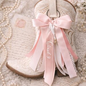 Pode incluir: Bota de cowboy marfim com bordados intrincados e detalhes em renda. Um grande la&ccedil;o rosa e fitas, uma com a inscri&ccedil;&atilde;o "Cloud 9", adornam a bota. A bota est&aacute; sobre um fundo vintage com p&eacute;rolas e notas manuscritas.