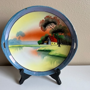 May include: A vintage blue and white porcelain plate with a hand-painted scene of a cottage by a lake with trees and a sunset. The plate has two handles on the sides.