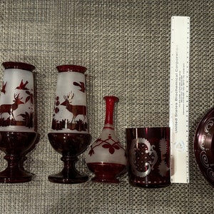 May include: A set of seven red glass etched vases and a lidded bowl. The vases and bowl feature white etched designs of flowers, birds, and deer. The bowl has a knob on the top.
