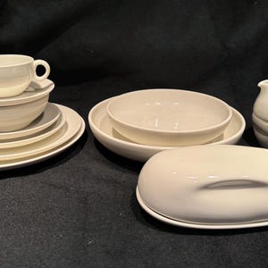 May include: A collection of white ceramic tableware, including plates, bowls, a cup and saucer, a creamer, and a covered butter dish. The items are arranged on a dark surface, showcasing a minimalist aesthetic.