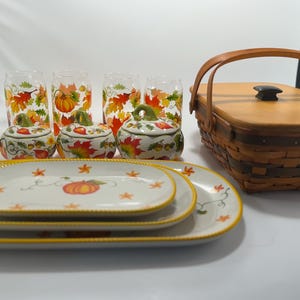 May include: A collection of fall-themed tableware and accessories. Includes clear glasses with pumpkin and leaf designs, small pumpkin-shaped lidded jars, oval plates with pumpkin and star designs, and a woven basket with a wooden lid and handle.