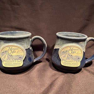 May include: Two blue ceramic mugs with a white glaze. Each mug has a gold oval sticker with the text "Hermann Hill Vineyard & Inn Hermann, Missouri www.hermannhill.com".