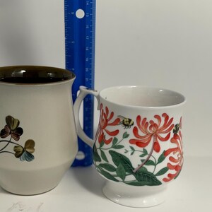 Puede incluir: Dos tazas de cer&aacute;mica. La taza de la izquierda es beige con un borde marr&oacute;n y un dise&ntilde;o floral. La taza de la derecha es blanca con un dise&ntilde;o floral en rojo y verde. Una regla azul est&aacute; entre las tazas.