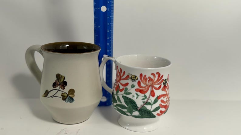 Puede incluir: Dos tazas de cer&aacute;mica. La taza de la izquierda es beige con un borde marr&oacute;n y un dise&ntilde;o floral. La taza de la derecha es blanca con un dise&ntilde;o floral en rojo y verde. Una regla azul est&aacute; entre las tazas.