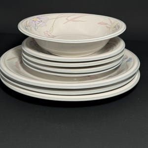 May include: A set of white ceramic dinner plates and a bowl with a floral design in shades of pink, purple, and green. The plates are stacked on top of each other, with the bowl on top of the stack.
