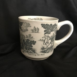 May include: White ceramic mug with a black and white design of horses and riders in a forest scene. The mug has the text "Catching the Mail" on the bottom.