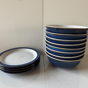 May include: A stack of eight blue bowls with a white interior and a thin brown rim. To the left, a stack of four matching plates with a white center and blue rim. The tableware is arranged on a white surface.