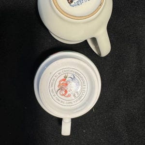 Puede incluir: Dos tazas de t&eacute; de cer&aacute;mica blanca. Una taza est&aacute; boca abajo, revelando un sello azul que dice "Designed in England". El interior de la otra taza muestra un dise&ntilde;o floral y el texto "Honeysuckle & Clematis".