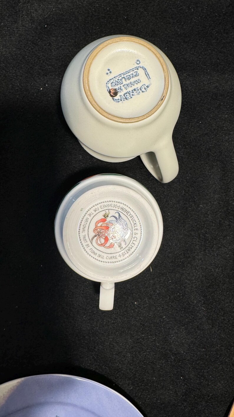 Puede incluir: Dos tazas de t&eacute; de cer&aacute;mica blanca. Una taza est&aacute; boca abajo, revelando un sello azul que dice "Designed in England". El interior de la otra taza muestra un dise&ntilde;o floral y el texto "Honeysuckle & Clematis".