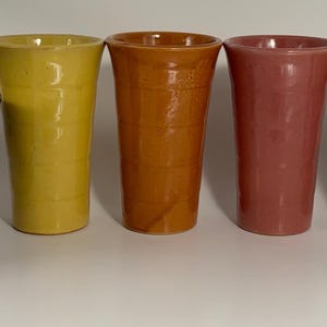 May include: A set of six ceramic cups in various colors, including yellow, orange, pink, beige, and green. The cups are all the same size and shape, and they are arranged in a row on a white surface. There is also a purple and brown ceramic bowl in the image.