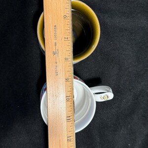 Puede incluir: Dos tazas de cer&aacute;mica con asas blancas y una regla de madera. Una taza es blanca con un dise&ntilde;o rojo y azul, la otra es marr&oacute;n. La regla indica que las tazas miden unos 10 cm de alto.
